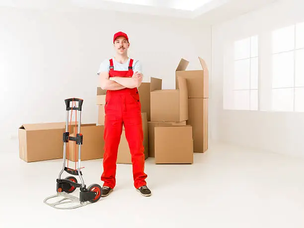 young confident delivery man standing in new home, holding hands crossed against his chest, with cardboard boxes in background