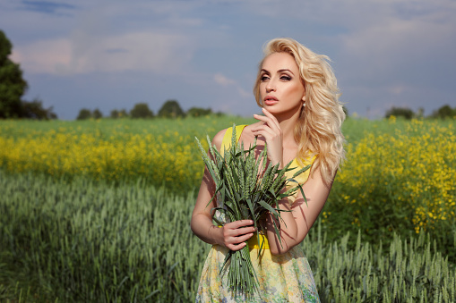 Girl stands in a field. In the background, yellow flowers. She's blonde.
