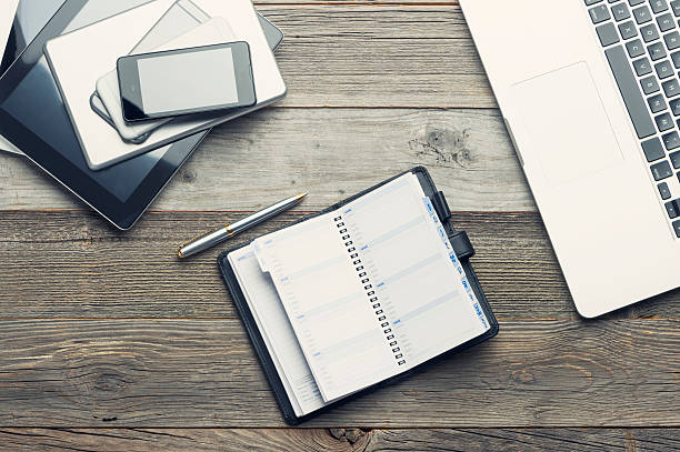 Stack of mobile devices and laptop on a wooden table. There are smart phones, digital tablets and mini digital tablets and phablets. 6 devices in all. All screens are blank. There is also part of a laptop keyboard. There is also a Filofax note pad with a pen. The wood is distressed an old. High angle view with copy space