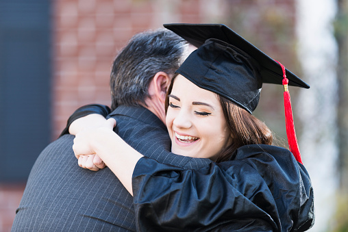 A teenage girl wearing a black graduation cap and gown, hugging her father, She is smiling, with her arms wrapped around her dad's neck, resting her chin on his shoulder, with her eyes closed.  We do not see his face so he is unrecognizable.  He is wearing a dark gray business suit.