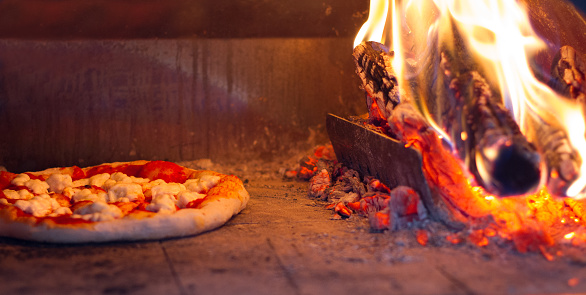 Baking pizza: looking into the inside of a pizza oven where a small pizza is cooking next to a wood fire.
