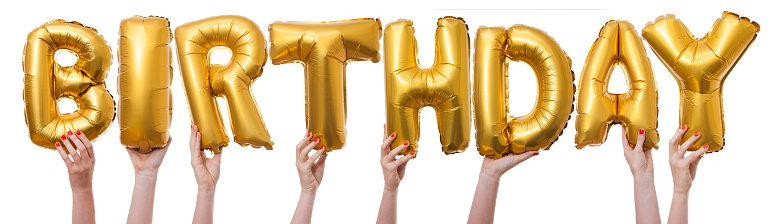 The word birthday has been created by female hands holding individual letter balloons. Each balloon is being held against a plain white background by a caucasian female hand. The Balloons are inflated and made from a shiny reflective gold material.