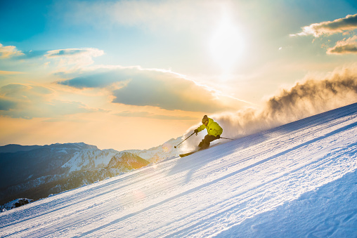 Man skiing and speeding down on ski slope at dusk, snowcapped mountain in background.