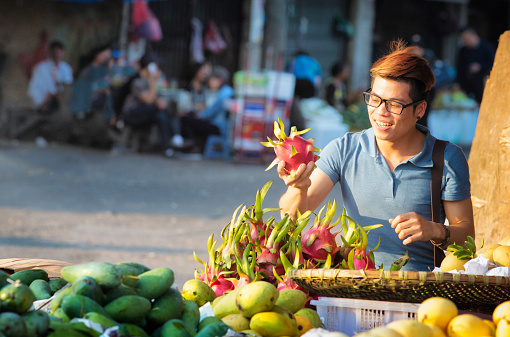 Young Vietnamese man picks up pitaya dragonfruit at Hanoi street market. He is smiling as he finds the best one. Lit by sun setting. Other fruits are desplayed around him.
