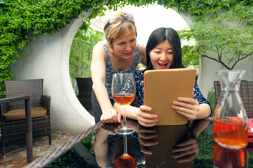 Mature blond caucasian woman and pretty young Chinese woman looking at a digital tablet in sandy brown cover. Young woman has long dark hair and dress with floral pattern. She is sitting at the table, the elderly woman is standing nearby.   Round door and blurred bamboo background. Outdoors, Europe. Nikon D3x, full frame, XXXL.