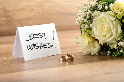 Close up Attractive Fresh Flowers, Pair of Rings and Best Wishes Card for Wedding on Top of Wooden Table.