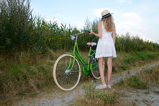 Teenage girl with long blond hair standing with green bike on a dirt road at a cornfield. The girl is wearing a white sundress, a sun hat and white sneakers. She has a northern european descent. Taken from rear view and low angle view. Sky with some light clouds on a summer evening in the background.