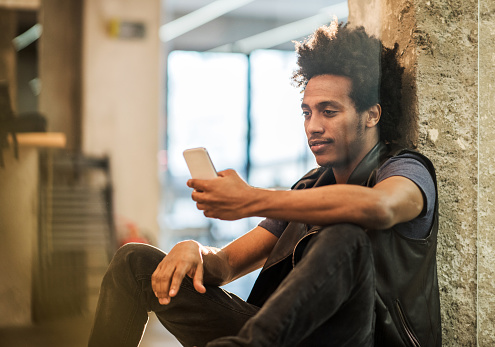 Smiling African American man sitting on the floor in the office and using cell phone. View is through the glass.