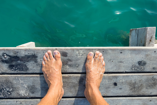 High angle POV of man's bare feet standing at edge of wooden dock sprinkled with fine white sand above aqua green sea water in natural morning light