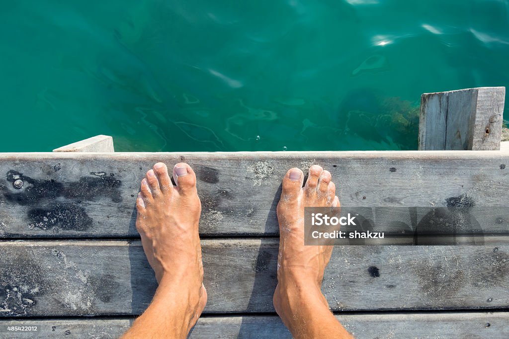 Ready to jump in High angle POV of man's bare feet standing at edge of wooden dock sprinkled with fine white sand above aqua green sea water in natural morning light Personal Perspective Stock Photo