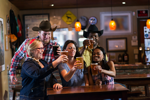 A multi-ethnic group of friends having fun at a restaurant bar, drinking beer.  Three women are sitting and two men are standing behind them, all raising their glasses in a celebratory toast.
