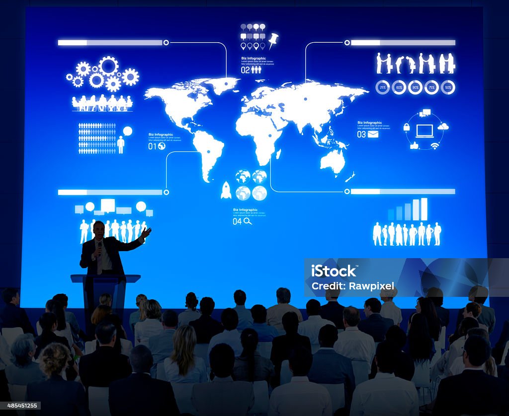 Instructor lectures to a crowd of businesspeople Business instructor standing in front of a blue screen, lecturing a group of people.  The blue screen features a map of the continents and various diagrams.  The people are seated in front of the instructor. Convention Center Stock Photo