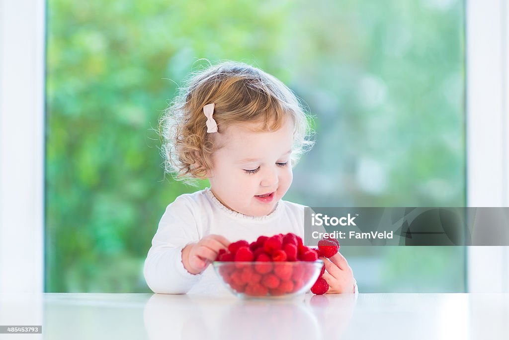 Happy smiling baby girl eating raspberry at white table Happy smiling baby girl eating raspberry at a white table next to a big window Baby - Human Age Stock Photo