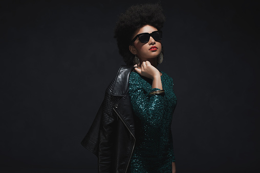 Half Body Shot of a Stylish Young Woman with Afro Hair, Holding her Jacket over the Shoulder against Dark Background.