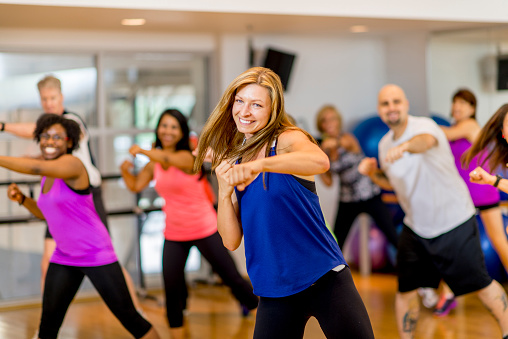 A woman is leading a multi-ethnic boxing class, everyone is wearing athletic clothing and is throwing a punch together - they are smiling and looking at the camera.