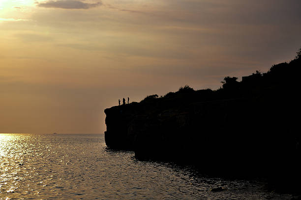Silhouettes of siblings standing in front of a stone pillar at sunrise.