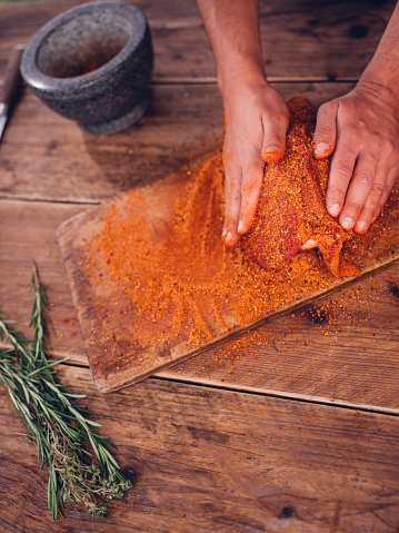 Spicy seasoning being rubbed into pork with herbs nearby