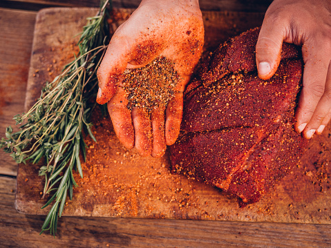 Overhead shot of a person's hand showing some spicy seasoning alongside a piece of quality raw pork on a vintage wooden table with herbs