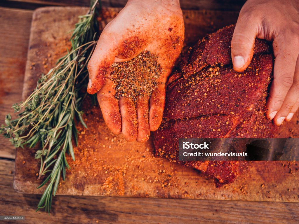 Hand showing spicy seasoning with a piece of raw pork Overhead shot of a person's hand showing some spicy seasoning alongside a piece of quality raw pork on a vintage wooden table with herbs Meat Stock Photo