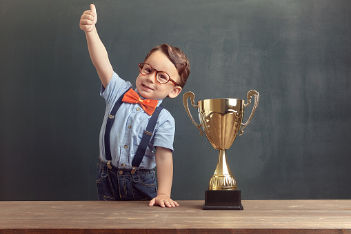 A cute, self-confident 2-3 years old boy is standing and showing his thumb up behind a wooden table with a golden trophy on it. Little boy is wearing an orange bow tie and blue trousers with suspenders.