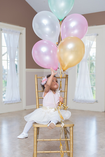 Preschool age girl in a lovely white tutu, sitting on a gold chair reaching for her party balloons.