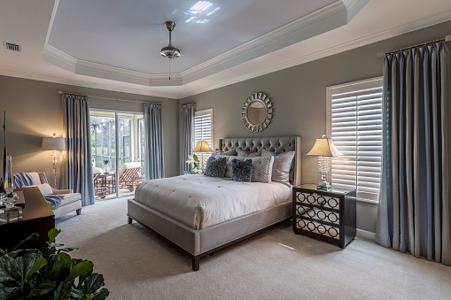 Large master bedroom interior in Southwest Florida home.  There is a large gray bed covered with a white bedspread.  There are furnishings and a green plant in the room, and the walls and curtains are gray.