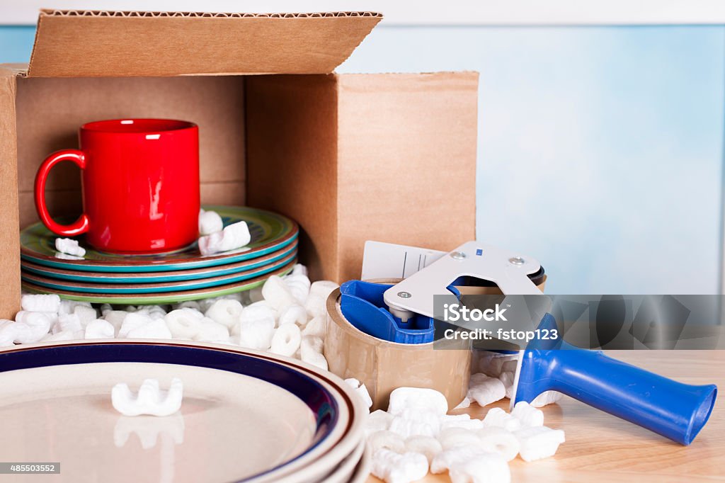 Moving. Dishes, cups packed inside cardboard box. Packing tape, peanuts. Relocation, moving concept. Plates, cups packed inside a cardboard box with white packing peanuts and a roll of packing tape on a blue dispenser.  The dishes are tucked inside the box to represent the concept of a family packing up to move.  A stack of plates is in foreground about to be packed.  2015 Stock Photo