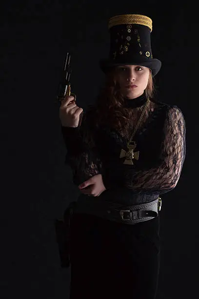 Cute brunette wearing a stovepipe hat and a Victorian-style dress, posing with a revolver and gunbelt - Steampunk fashion.