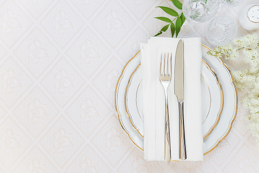 Top view of the beautifully decorated table with white plates, crystal glasses, linen napkin, cutlery and white flower on luxurious tablecloths, with space for text