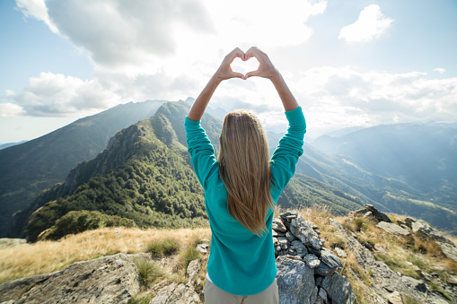 Young woman hiker on the mountain top making a heart shape with her hands above her head showing she is loving the nature.