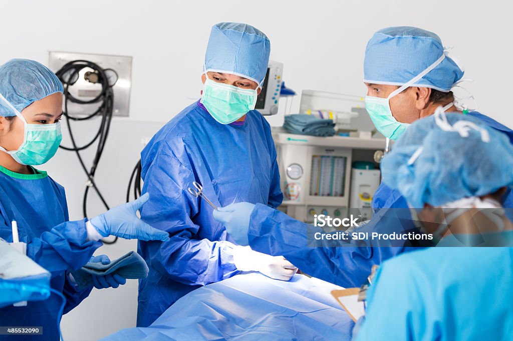 Surgeon handing instruments to technician or nurse during operation Maturea adult Caucasian doctor is handing clamp or surgical instrument to young adult Asian intern or surgical nurse during operation in hospital. Team of surgeons is operating on patient. Doctors are wearing surgical gowns, caps, masks and gloves. Giving Stock Photo