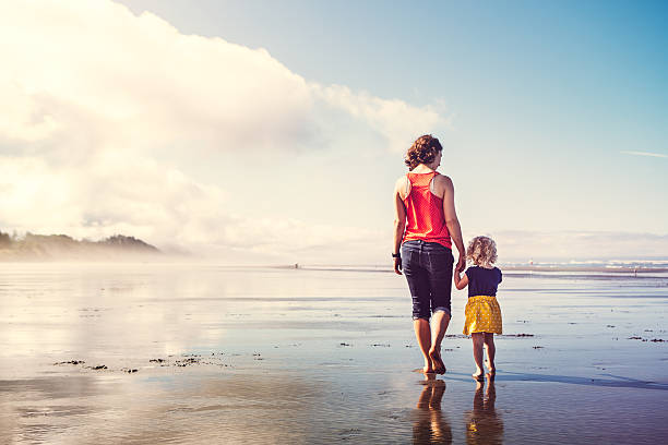 Beach Girls Walking Hand in Hand A young woman and a little girl (mother / daughter or nanny / caretaker) walk on a beautiful sunny Washington state beach holding hands, their reflections visible in the wet sand.  A stunning vacation landscape showing the Pacific ocean coast during summer weather.  Horizontal with copy space, the women walking away from the camera. washington state coast stock pictures, royalty-free photos & images