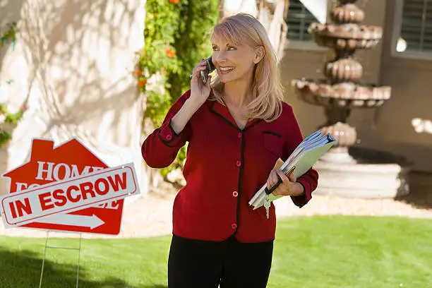 Happy female real estate agent with files answering mobilephone in front of house