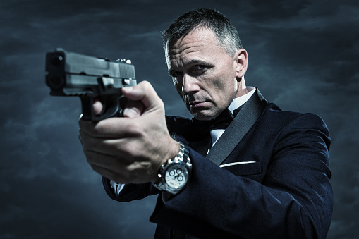 A handsome mature male secret agent, bodyguard, spy, or security staff dressed in an elegant tuxedo and bow tie points a gun on a stormy night with cloudy sky in the background.