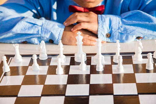 A Latin or Indian descent little boy plays chess outdoors. He has a serious expression as he studies the chess board in preparation for his next move. He wears a blue long-sleeve shirt and red bow tie. 