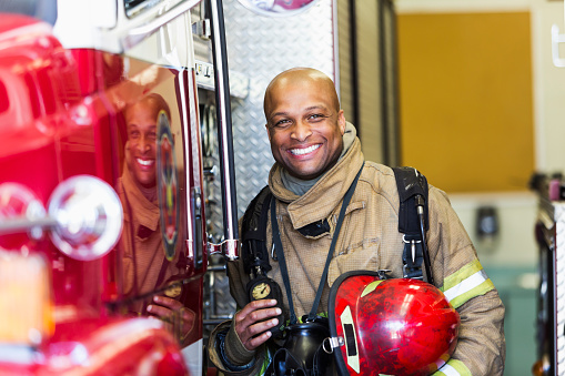 Portrait of an African American firefighter standing next to a fire truck.  He is wearing a protective suit and holding a red helmet.  He is looking at the camera, smiling.