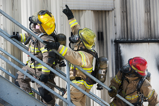 A group of three multiracial firefighters, led by a woman, climbing up a metal staircase on the exterior of an industrial building or warehouse.  They are wearing protective clothing, helmets and oxygen masks.  The man in the middle is pointing upward.