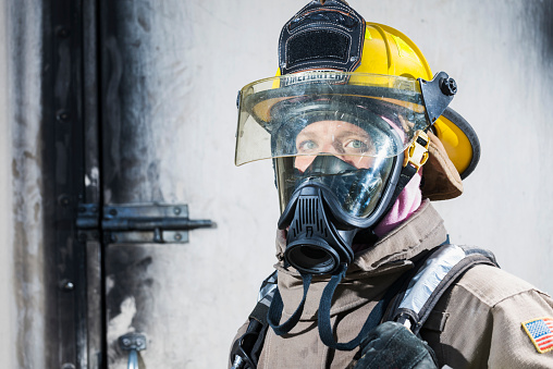 The face of a female firefighter, wearing a protective suit, helmet and oxygen mask.  Only her eyes are visible underneath the gear.