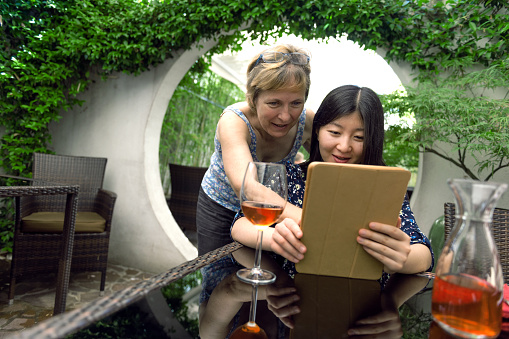 Mature blond caucasian woman and pretty young Chinese woman looking at a digital tablet in sandy brown cover. Young woman has long dark hair and dress with floral pattern. She is sitting at the table, the elderly woman is standing nearby.   Round door and blurred bamboo background. Outdoors, Europe. Nikon D3x, full frame, XXXL.