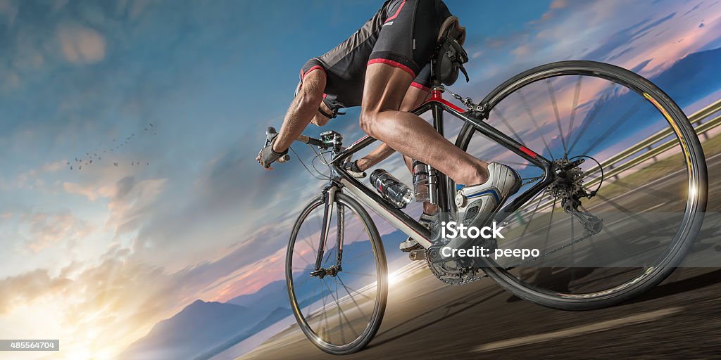 Cyclist Moving Fast On Coastal Path An image of a professional Caucasian male cyclist / triathlete dressed in cycling shorts, top, cycling shoes and safety helmet, riding a racing bike at high speed along a coastal road close to sea and mountains under a dramatic evening sky at sunset.  With motion blur on the road and wheels.  Cycling Stock Photo