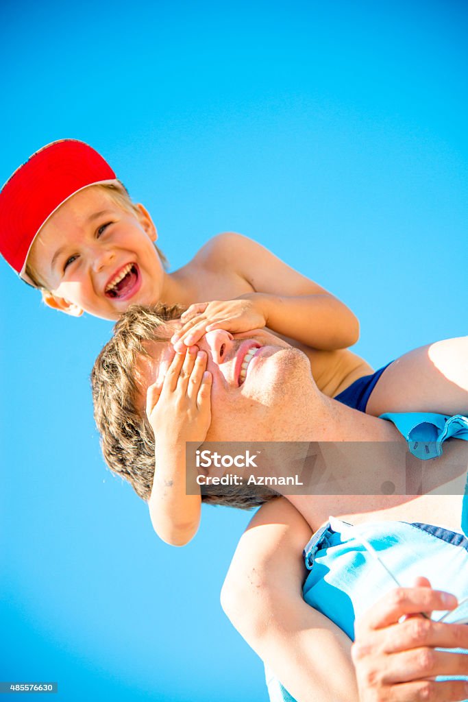 Enjoying Summer Little boy sitting on his father's shoulders and he is covering father's eyes with his hands. He is smiling. 2015 Stock Photo