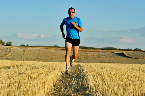 young sport man with sunglasses running outdoors on straw field ground in frontal perspective towards camera in healthy lifestyle and summer training concept