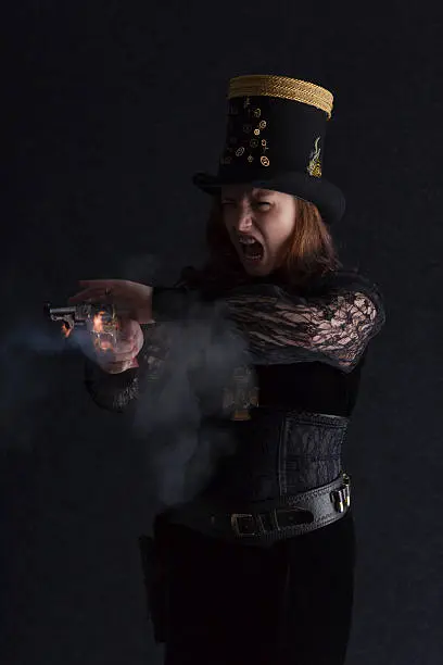 Focus on the shootist in this image of a cute brunette wearing a stovepipe hat and a Victorian-style dress - Steampunk fashion. Camera activated as the revolver is fired to get genuine flame and gunsmoke!