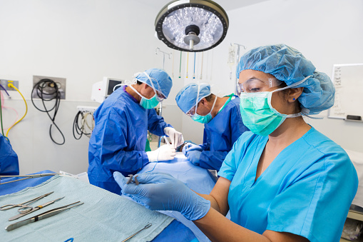 Mid adult Hispanic female nurse or surgical technician is prepping instruments for surgeons. Surgeons are performing surgery on patient in background. Modern operating room has halogen lights and newest technology. Doctors are wearing surgical gowns, caps, gowns, gloves and masks.