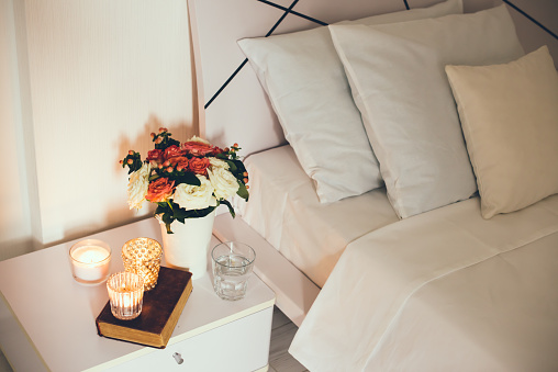 Interior of white bedroom, new linens on the bed, the room at the hotel. Bedside table decor with candles and pillows closeup.