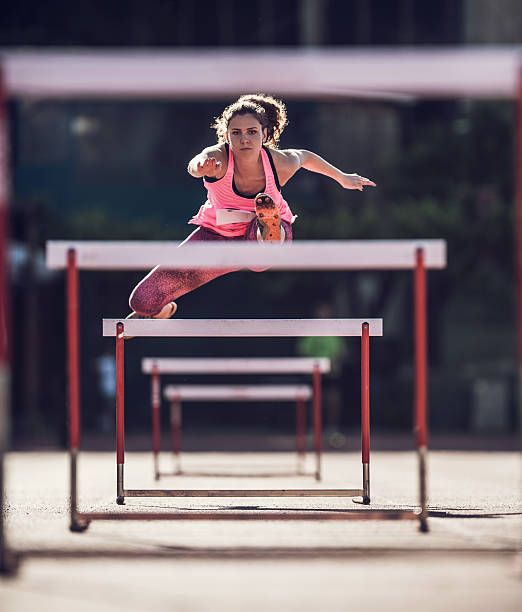 A female collegiate heptathlete powers over a hurdle at full speed during an intense solo training session with her coach observing nearby.
