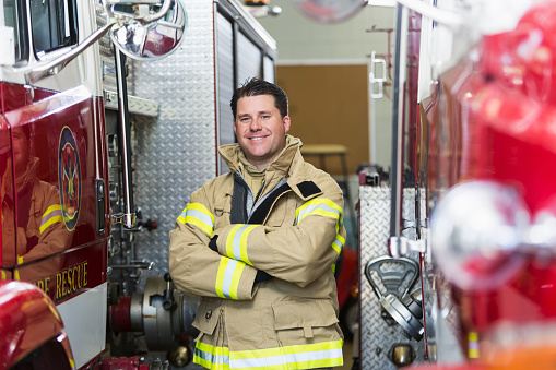 Portrait of a fireman wearing his fire fighter suit, standing with arms folded next to fire engine at the station.  He is looking at the camera with a serious expression.