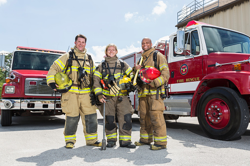 Outdoor portrait of diverse female and male firefighters posing in front of the fire engine, looking at the camera, holding their helmets.