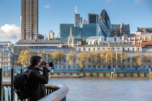 Man Photographer in London, UK. The man is taking picture close to the Thames River with the cityscape in the background.