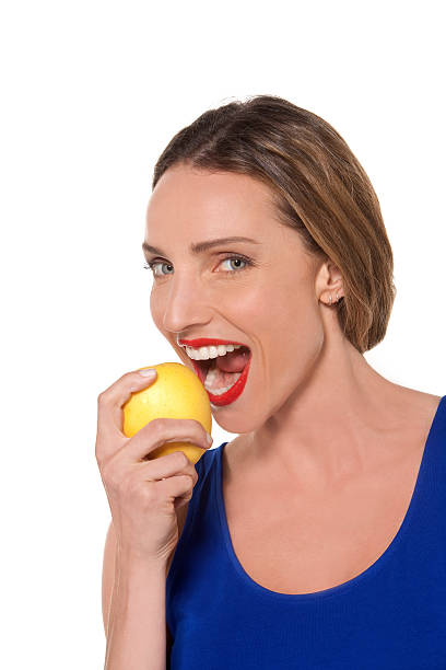 Eating an apple is good for you Portrait of a woman wearing a blue top eating a yellow apple. She has red lipstick too. Studio shot isolated on white with copy space. apple women smiling human teeth stock pictures, royalty-free photos & images